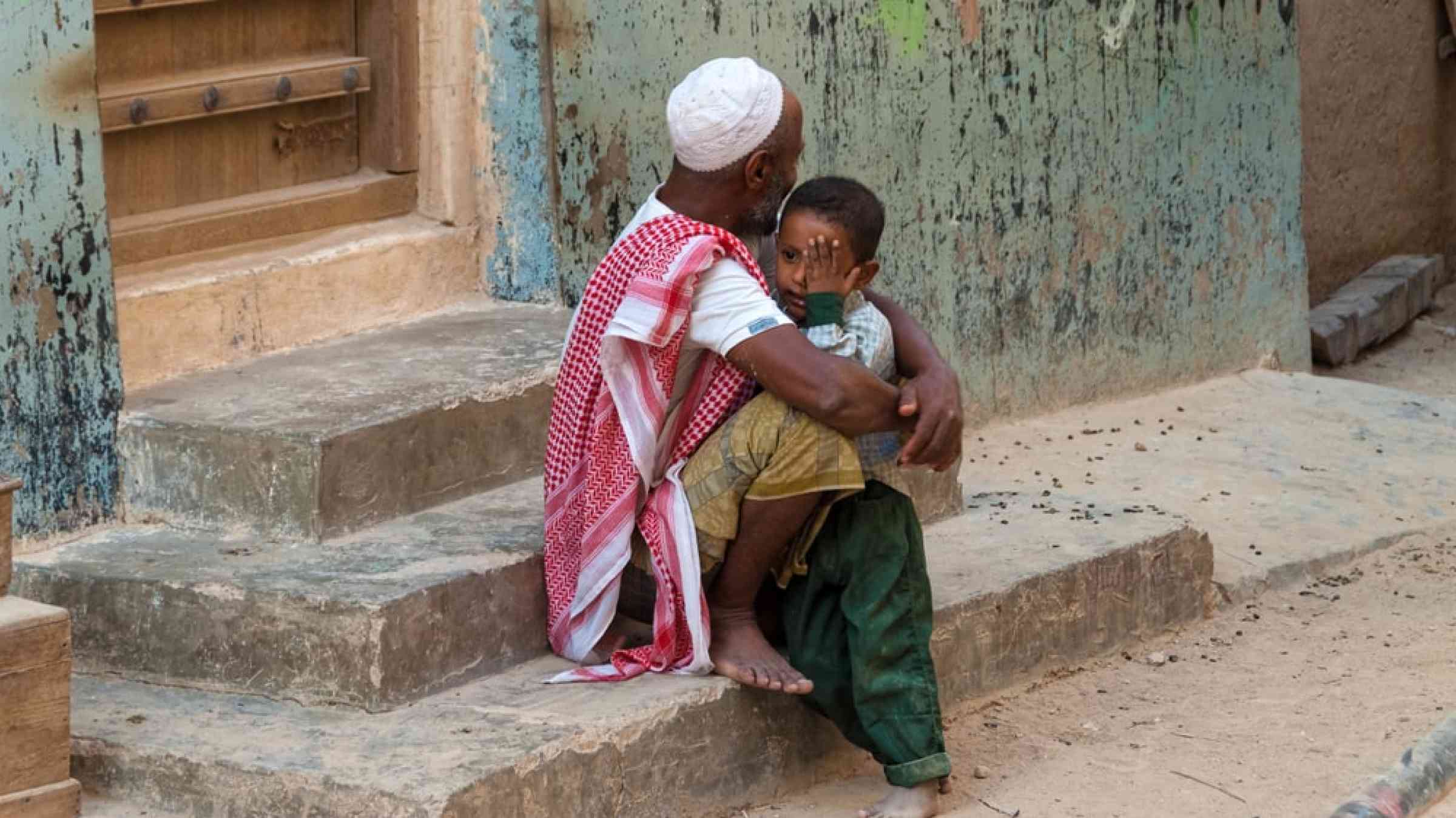 A farther holds his son in his arms while sitting on the stairs in front of his house in Yemen.