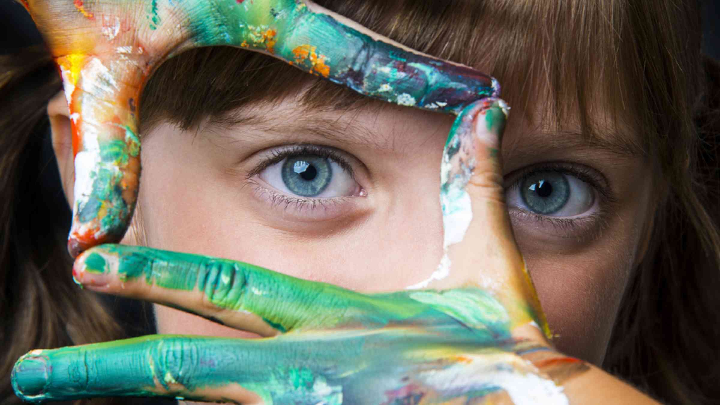 A small girl with colored hands in front of her face.