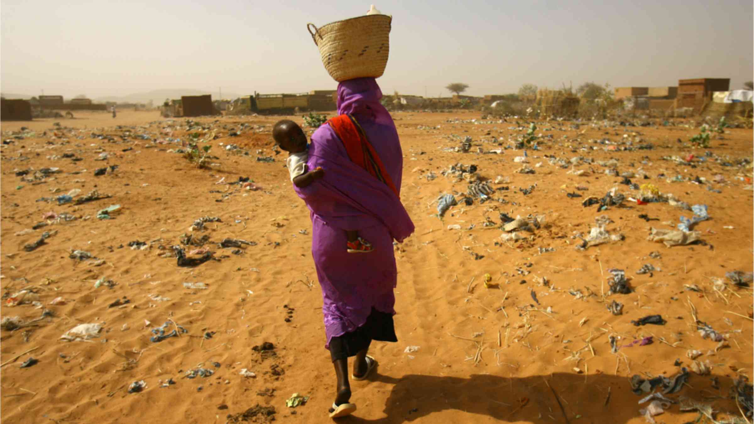 South Sudanese woman carrying her baby through polluted sand field.